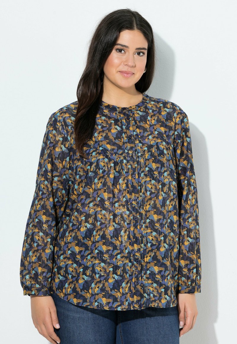Frau mit langen dunklen Haaren, die eine blau, orange und schwarz gemusterte Bluse mit Knöpfen und blaue Jeans trägt, steht vor einer weißen Wand.