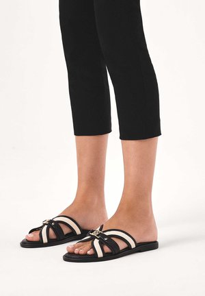 Pieds en sandales plates noires et beige à glissière avec boucle décorative, portant un pantalon noir raccourci sur un fond blanc uni.