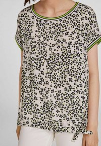 Let blouse i creme med sort og grøn blomsterprint. Har rund halsudskæring, korte ærmer og en knyttet detalje på siden.