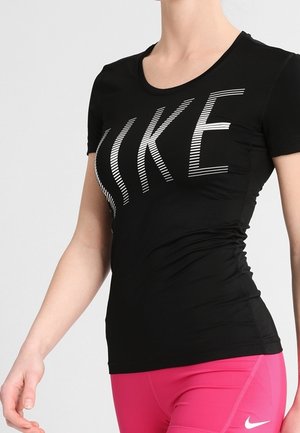 Personne portant un t-shirt noir Nike avec un logo blanc et un short Nike rose vif, debout devant un fond clair uni.