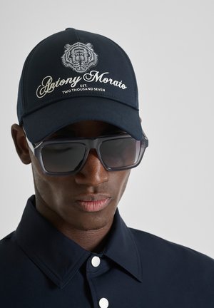 Muškarac koji nosi crnu kapu s bijelim logotipom tigra i tekstom "Antony Morato", velike tamne sunčane naočale i tamnoplavu košulju s ovratnikom.