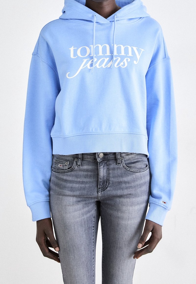 Sudadera corta de color azul claro con el logo blanco de "tommy jeans", material de algodón, capucha con cordón y puños acanalados; combinada con jeans ajustados grises.