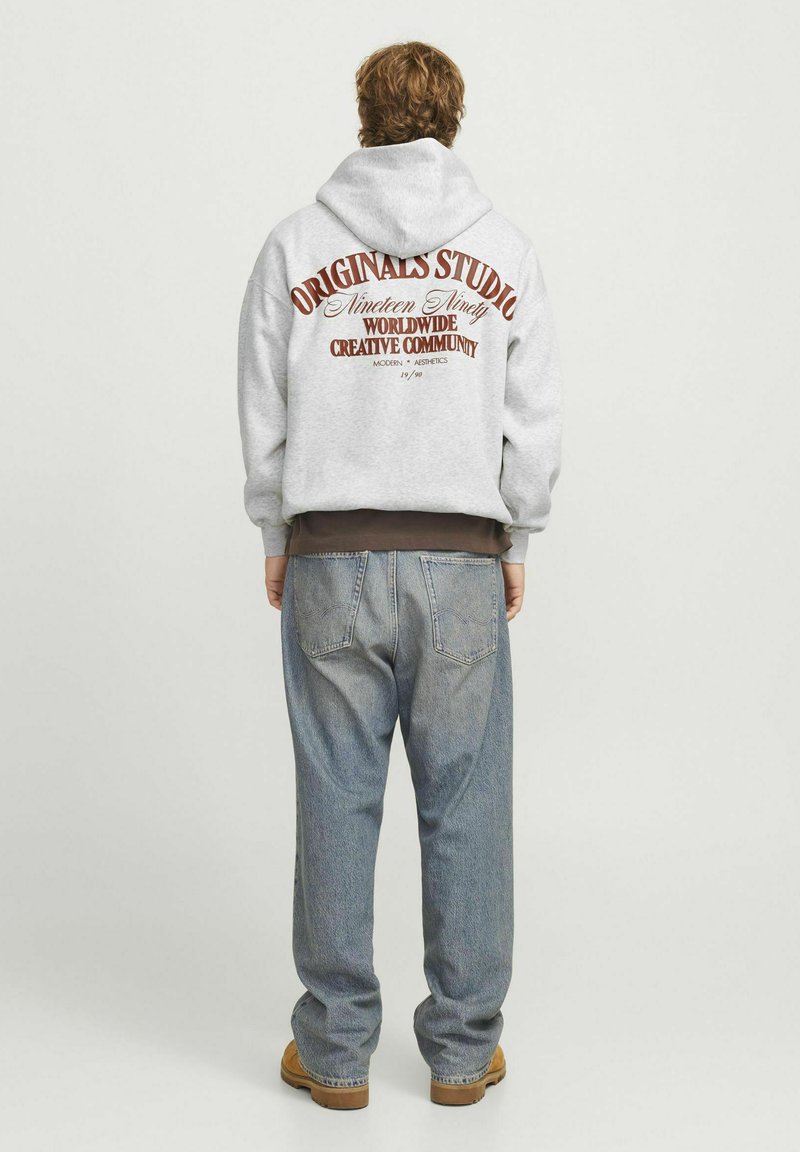 Sudadera gris con un gran logotipo impreso en la parte posterior. La persona lleva unos pantalones vaqueros holgados y botas marrones. El material parece suave y acogedor.