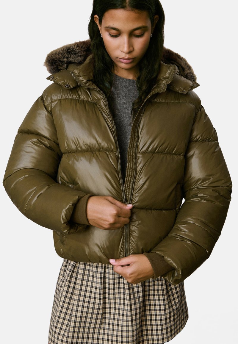 Une femme portant une veste matelassée marron avec une capuche en fourrure, un pull gris et une jupe à carreaux, ferme la fermeture éclair de sa veste tout en regardant vers le bas.