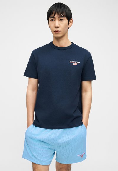 Άνδρας που φοράει μπλε σκούρο μπλουζάκι Polo Sport και ανοιχτό μπλε σορτς από δίχτυ, με τα χέρια στις τσέπες, στέκεται μπροστά σε απλό λευκό φόντο.