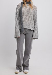 Femme portant un pull gris à mailles ouvertes, une écharpe assortie, un jean gris à jambes larges et des baskets blanches, debout devant un fond uni.