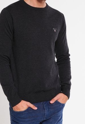 Suéter de punto gris oscuro con mangas largas, puños y dobladillo acanalados, que presenta un pequeño logo en el pecho. Combinado con jeans azules.