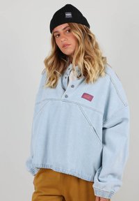 Pull en denim bleu clair avec col boutonné, poignets ajustables et un petit patch logo bordeaux. Présente une coupe décontractée et oversize.