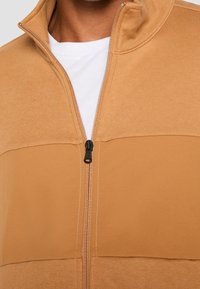 Giacca con zip color camoscio caratterizzata da un design bicolore, tessuto morbido, colletto e dettaglio della zip nera. Include uno strato interno bianco.