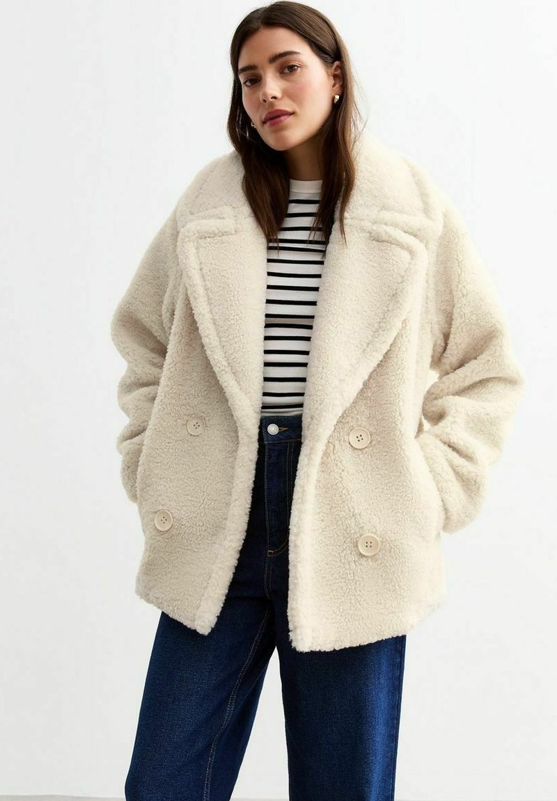 Crèmefärgad imitationpälsjacka med dubbelknäppning, breda slag och stora knappar, som kombineras med en randig skjorta och mörk denimjeans.