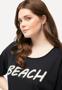 Sort t-shirt med afslappet pasform, med hvide fluffy bogstaver der staver "BEACH" på forsiden. Langt bølget brunt hår, sølvfarvede hoops.