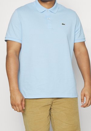 Homme portant un polo à manches courtes bleu clair avec un petit logo crocodile vert et un pantalon beige, debout devant un fond uni.