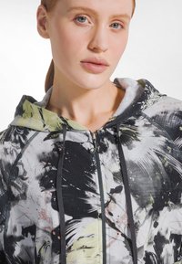 Joven mujer de piel clara y ojos azules que lleva una sudadera con capucha con cierre, estampada con un patrón abstracto en negro, blanco y verde, con capucha y cordones.