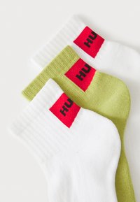 Trois paires de chaussettes : une blanche, une verte, et une rayée blanche, chacune avec un bandeau rouge portant du texte noir au niveau de la cheville.