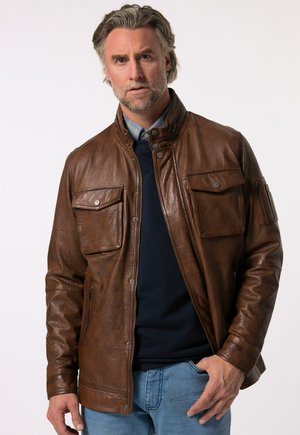 Braune Lederjacke mit strukturiertem Finish, ausgestattet mit zwei Brusttaschen, einem hohen Kragen und einem Reißverschluss vorne. Getragen über einem marineblauen Pullover.