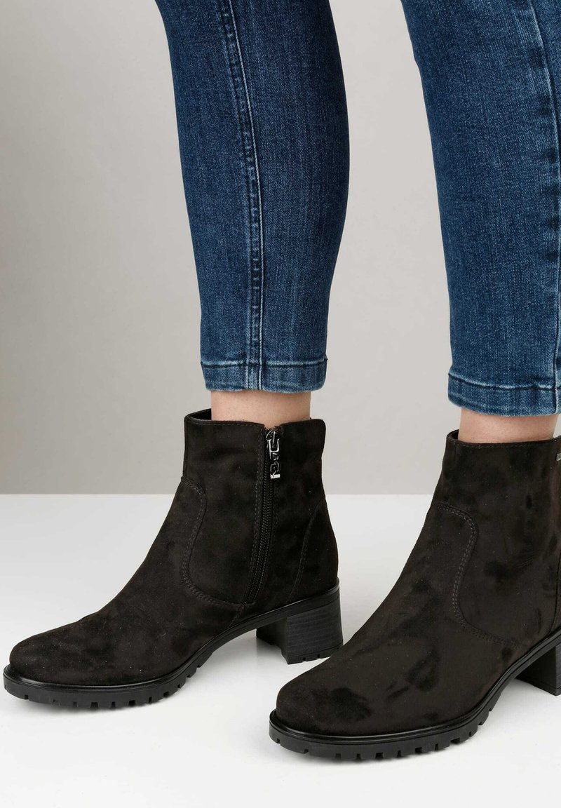 Botines de ante negros con tacón cuadrado y cierre de cremallera. Presentan una textura suave y punta redonda, combinados con jeans ajustados azules.