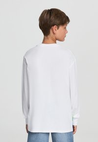 Biała koszula z długim rękawem o luźnym kroju, z okrągłym dekoltem i subtelnym graficznym detalem na ramieniu; zestawiona z niebieskimi dżinsami.