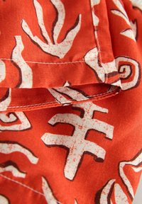 Czerwona tkanina z abstrakcyjnymi białymi i brązowymi wzorami, z widocznym przeszyciem i fakturą. Wzór zawiera wiry i kształty.