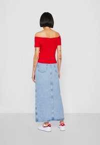 Rød off-shoulder top parret med en lys blå denim maxi nederdel, der har to baglommer og frynset kant. Båret med hvide sneakers.