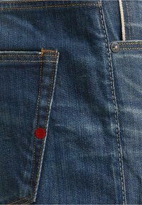 Tela de mezclilla en azul oscuro con costuras doradas, que presenta un bolsillo de parche y un pequeño acento rojo cerca de la costura, mostrando un diseño clásico.