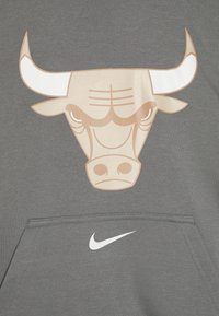 Sivá majica s kapuco z velikim grafičnim motivom bika v bež barvi z belimi poudarki. Vključuje sprednji žep in bel Nike swoosh logotip.