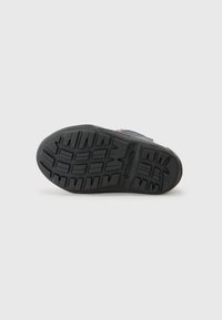 Semelle de chaussure noire avec un motif texturé et un logo, sur un fond blanc uni.