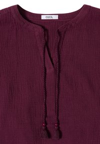 Blouse bordeaux texturée à manches longues avec décolleté fendu, liens en tresse à pompons avec perles, et étiquette intérieure indiquant "CECIL".