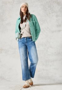 Türkiser Fleece-Jacke, beiger Strickpullover, hellblaue Jeans und beiger Sneaker. Kleine mintgrüne Umhängetasche. Sanfte Texturen, lockere Passform.