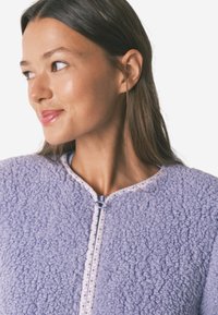 Polarowa kurtka w jasnym fioletowym kolorze z fakturowaną powierzchnią, zamek błyskawiczny oraz kontrastowe wzorzyste wykończenie wzdłuż dekoltu i zamka.