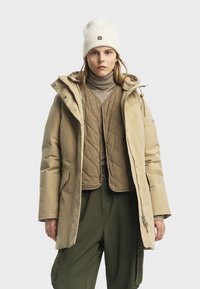 Giacca puffer beige con cappuccio, dotata di uno strato interno trapuntato, chiusura con zip e grandi tasche, abbinata a un dolcevita beige e pantaloni verdi.