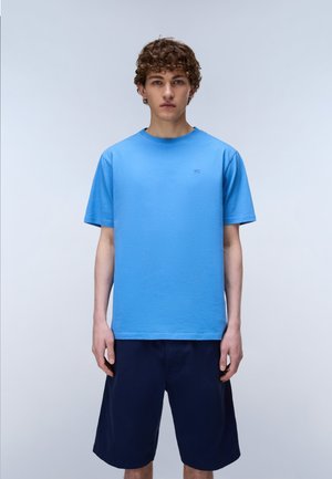 Junger Mann steht, trägt ein schlichtes hellblaues kurzärmeliges T-Shirt und dunkelblaue knielange Shorts vor einem einfarbigen Hintergrund.