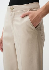 Pantalons beige à jambes larges avec poches latérales, fermeture à bouton et texture lisse et légère. Coupe ajustée à la taille.