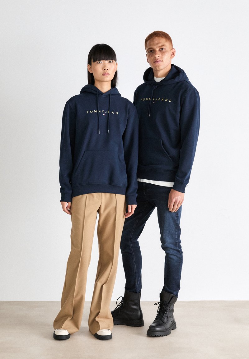Sweatshirts bleu marine avec le logo doré "TOMMY JEAN" ; le modèle porte un pantalon beige, son partenaire en jeans foncés et bottes noires. Tissu lisse, design décontracté.