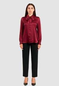 Blusa in satin bordeaux con colletto a fiocco, maniche lunghe e chiusura con bottoni. Abbinata a pantaloni neri su misura e tacchi neri.
