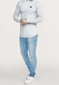 Szara koszula z długim rękawem o dopasowanym kroju z małym czarnym logo na piersi. Noszona z jasnoniebieskimi jeansami o dopasowanym kroju i białymi trampkami.