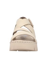 Sandales en cuir beige avec des straps croisées, un bouton circulaire décoratif et une semelle intérieure texturée. Semelle épaisse avec un léger relief.