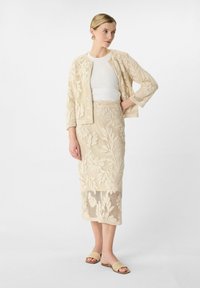 Beige Spitzenjacke mit floralem Muster, kombiniert mit einem passenden knielangen Rock und einem weißen Oberteil. Braune Rutsch-Sandalen runden das Outfit ab.