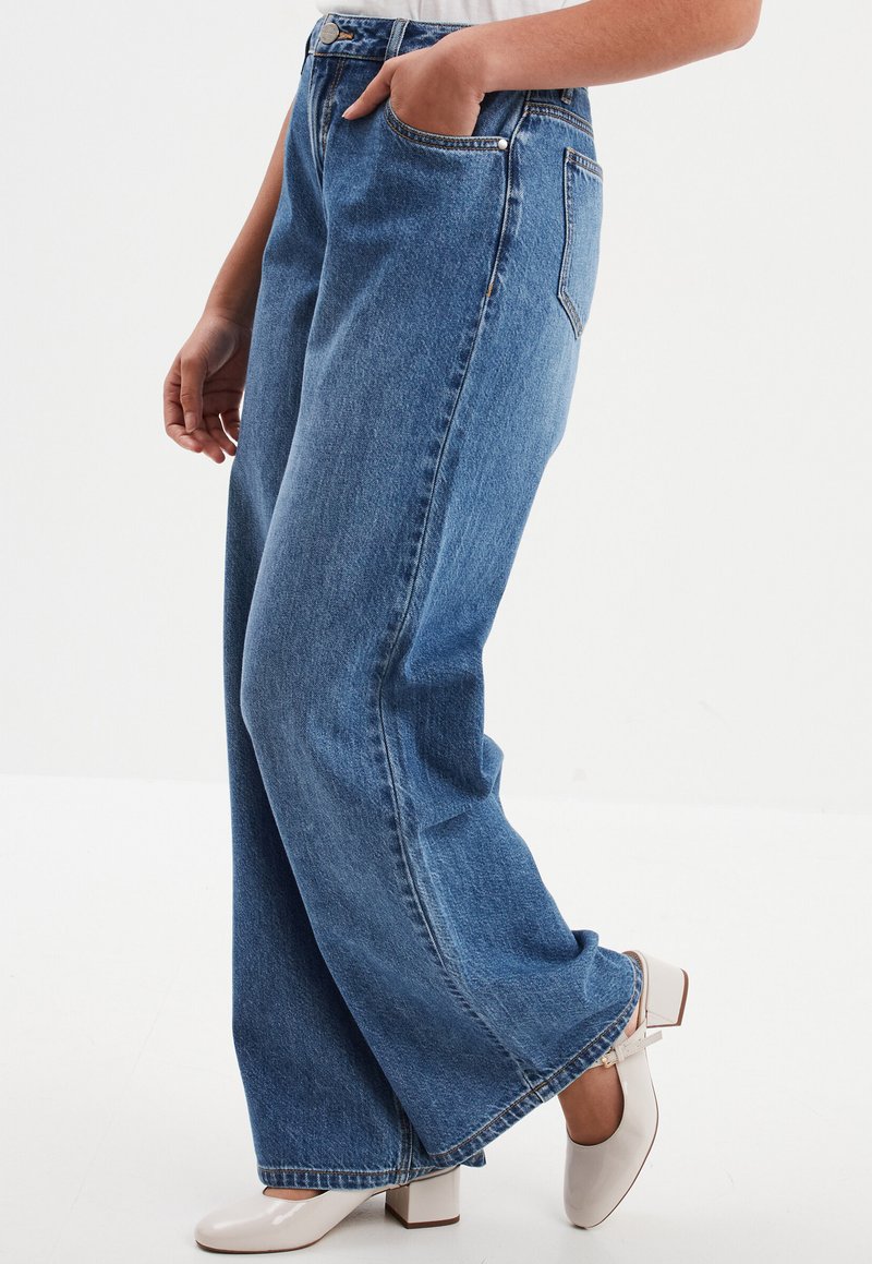 Hochgeschnittene, weit geschnittene Jeans aus dunkelblauem Denim mit glatter Textur. Verfügt über Seitentaschen und einen cleanen Saum. Kombiniert mit weißen Absätzen.