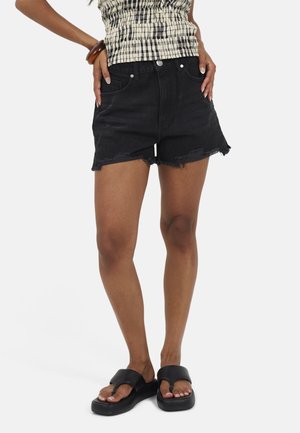 Donna che indossa pantaloncini di jeans neri usurati, sandali con plateau neri e una maglietta fantasia con un braccialetto al polso destro.