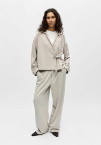 Femme aux longs cheveux bouclés portant une veste beige ample nouée à la taille, un pantalon large assorti et des chaussures noires à bouts pointus.