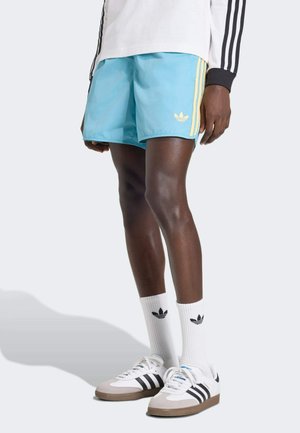 Pantalones cortos de baño azul claro con rayas laterales amarillas y un logo de Adidas. Combinados con calcetines blancos hasta la pantorrilla con un logo negro y zapatillas en blanco y negro.