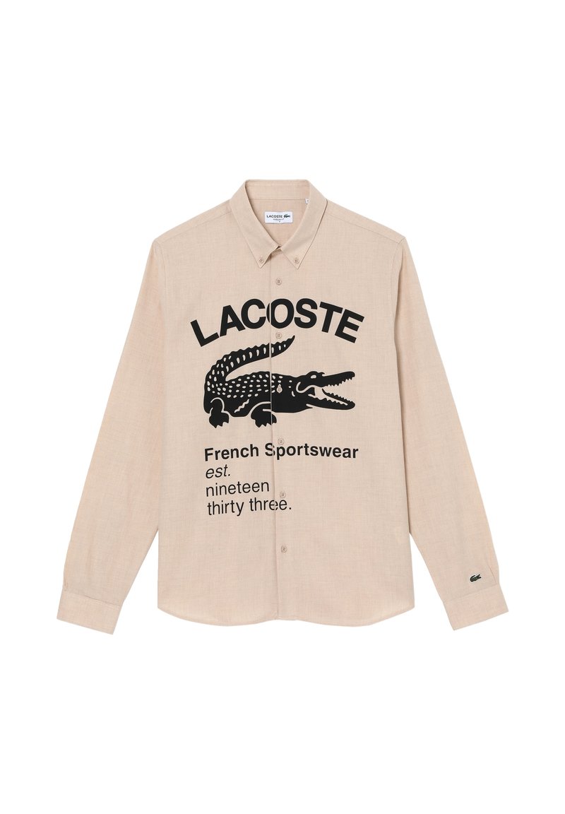 Chemise beige à manches longues avec boutons, illustrée d'un crocodile noir, de texte audacieux et d'un petit logo vert sur le poignet. Tissu en mélange de coton.