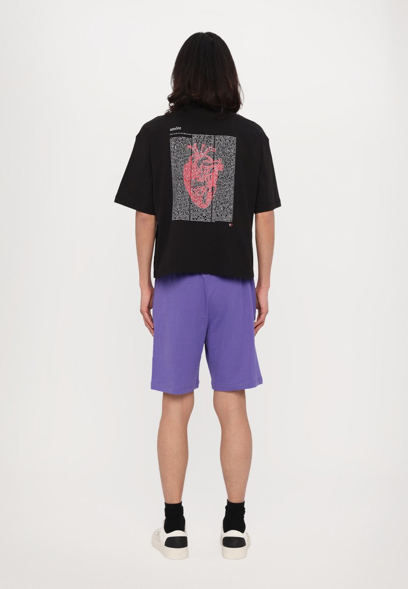 Juoda trumpa rankovė t-shirt su raudonos širdies grafika ir tekstu ant nugaros, derinama su violetiniais šortais ir baltais sportbačiais.