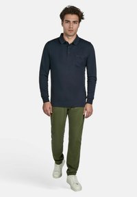 Polo à manches longues bleu marine avec un col et une poche poitrine, associé à un pantalon slim-fit vert olive et des baskets blanches.