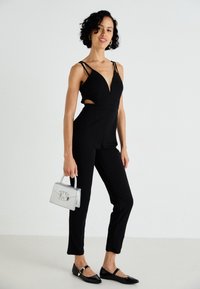 Tuta nera con scollatura profonda e lati sagomati. Abbinata a una borsa argentata e ballerine nere. Tessuto liscio, design aderente.