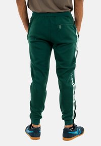 Joggers de color verde oscuro con un ajuste tapered, que presentan rayas laterales blancas, una cintura elástica y detalles en el bolsillo trasero. Combinados con zapatos oscuros.