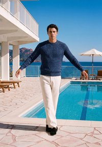 Homme portant un pull bleu et un pantalon blanc marchant à côté d'une piscine avec des chaises longues, un parasol et une vue sur l'océan en arrière-plan.