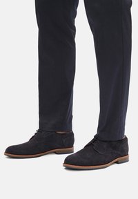 Czarne zamszowe botki za kostkę z wiązanym przodem i brązową podeszwą. Posiadają okrągły nosek i subtelne akcenty w postaci przeszyć. W zestawie z ciemnymi spodniami.