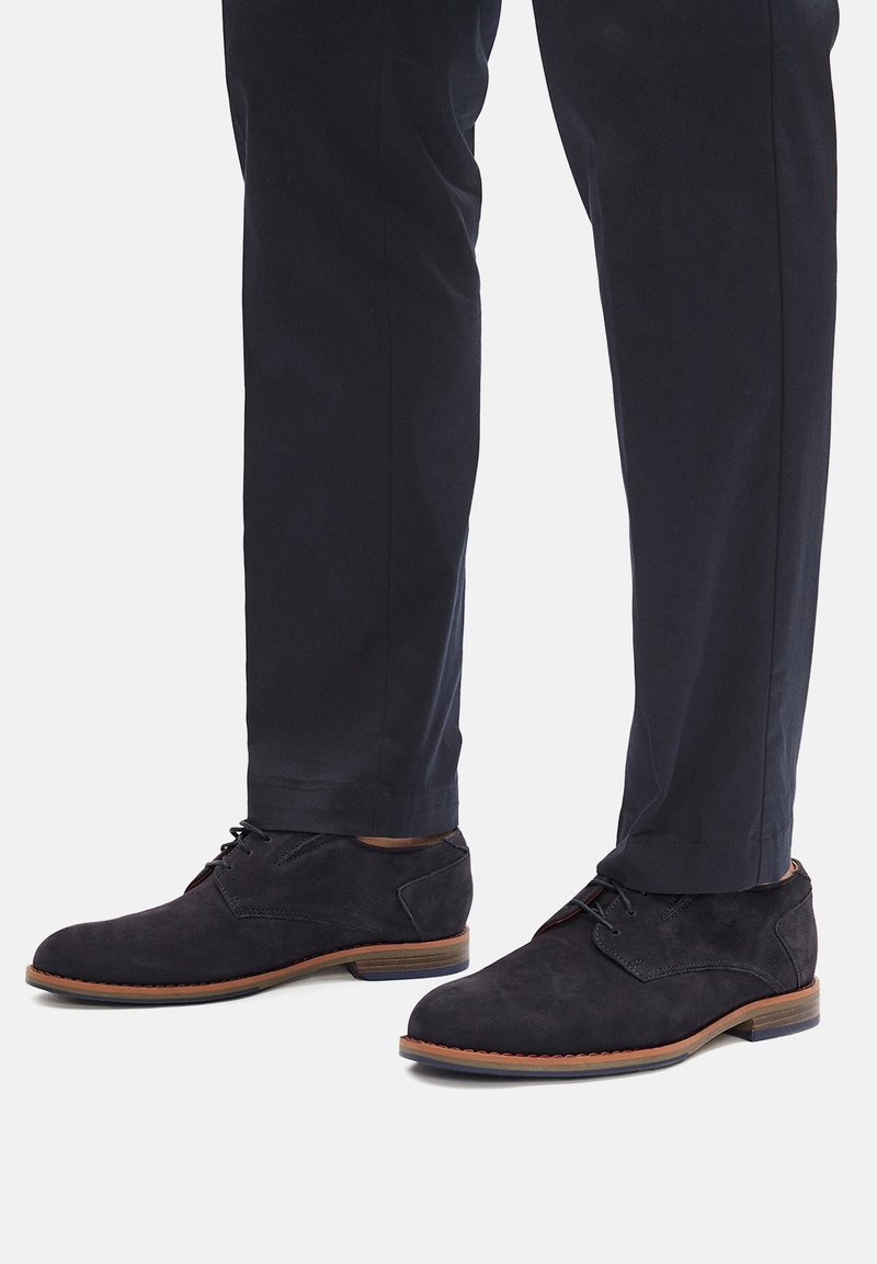 Czarne zamszowe botki za kostkę z wiązanym przodem i brązową podeszwą. Posiadają okrągły nosek i subtelne akcenty w postaci przeszyć. W zestawie z ciemnymi spodniami.