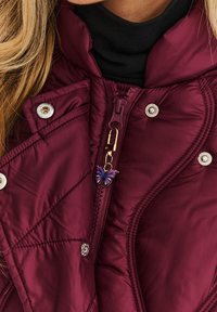 Bordová prešívaná bunda s vysokým golierom, so zlatým zipsom ozdobeným príveskom vo forme motýľa a striebornými cvokmi.